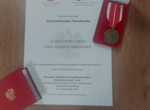 Na drewnianym blacie leży zaproszenie na uroczystość z okazji Dnia Edukacji Narodowej, skierowane do Pani Aleksandry Twardowskiej, oraz medal Komisji Edukacji Narodowej w czerwonym etui.