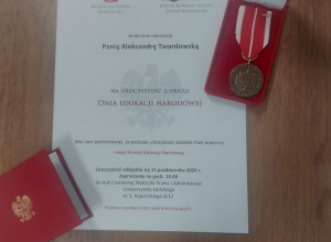 Na drewnianym blacie leży zaproszenie na uroczystość z okazji Dnia Edukacji Narodowej, skierowane do Pani Aleksandry Twardowskiej, oraz medal Komisji Edukacji Narodowej w czerwonym etui.