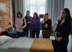 grupa uczennic stoi wokół pojedynczego łóżka w jednostce mieszkalnej typu SINGLE i słuchają wypowiedzi osoby oprowadzającej na temat wyposażenia jednostki mieszkalnej