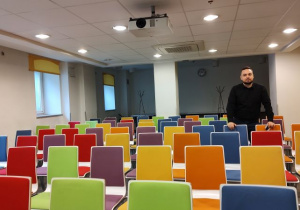kierownik operacyjny hotelu stoi wśród kolorowych krzeseł w Sali konferencyjnej i opowiada o przebiegu konferencji