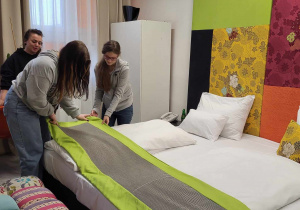 Uczniowe ścielą łóżko w pracowni hotelarskiej pod okiem pracownika hotelu